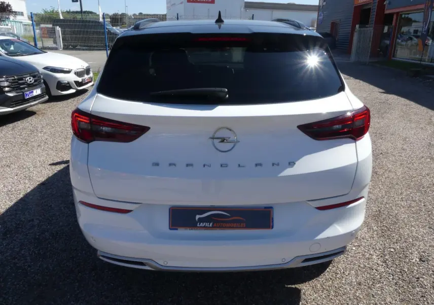 Vue arrière d'un Opel Grandland blanc métallisé avec vitres arrière surteintées et feux LED, en extérieur sur parking.
