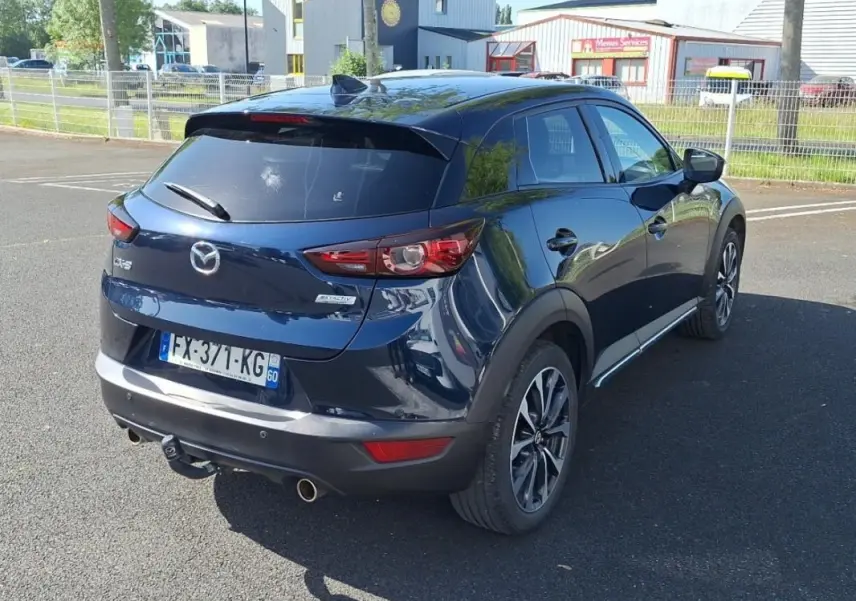 Vue 3/4 arrière droite d'un Mazda CX-3 bleu avec jantes aluminium et attelage visible sur parking.