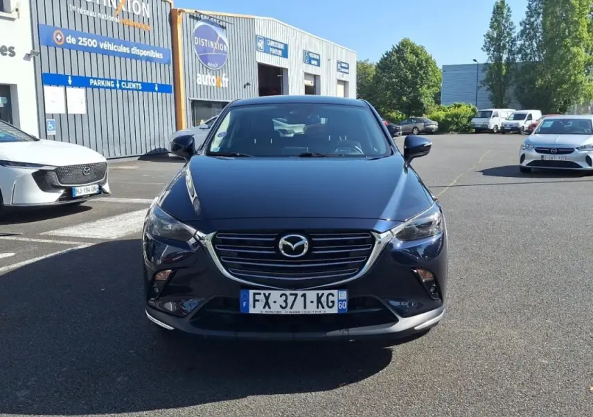 Vue avant d'un Mazda CX-3 bleu foncé garé devant un garage, avec calandre chromée et phares allumés.