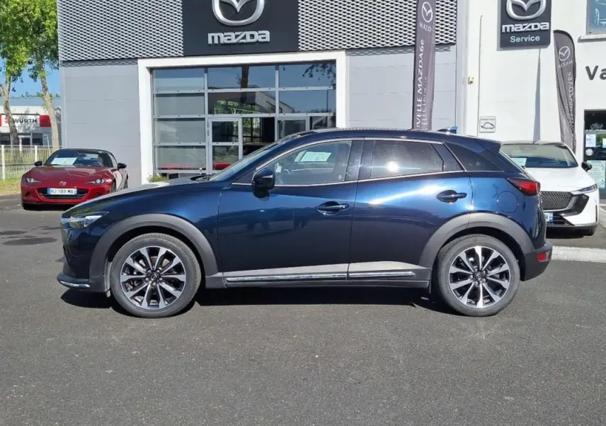 Vue de profil côté gauche d'un Mazda CX-3 bleu 2021 stationné devant un concessionnaire Mazda.