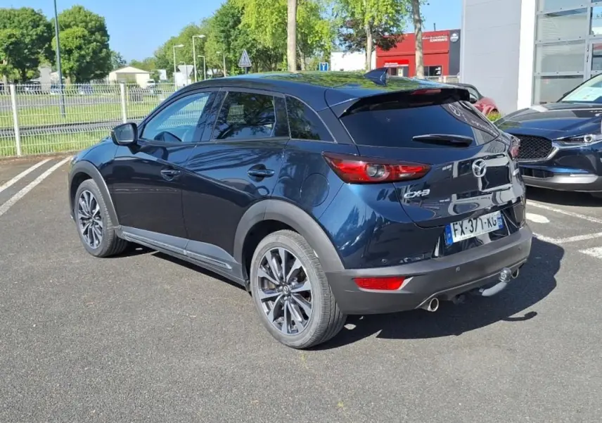 Vue 3/4 arrière droite d'un Mazda CX-3 bleu 2021 avec jantes aluminium et attelage visible en extérieur.