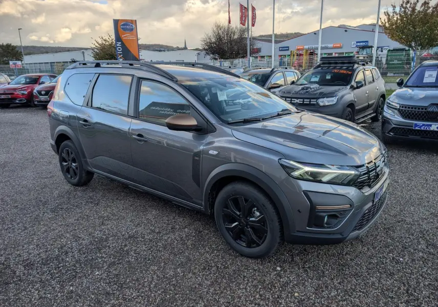 Dacia Jogger gris métallisé en 3/4 avant droit avec jantes noires et barres de toit sur parking.