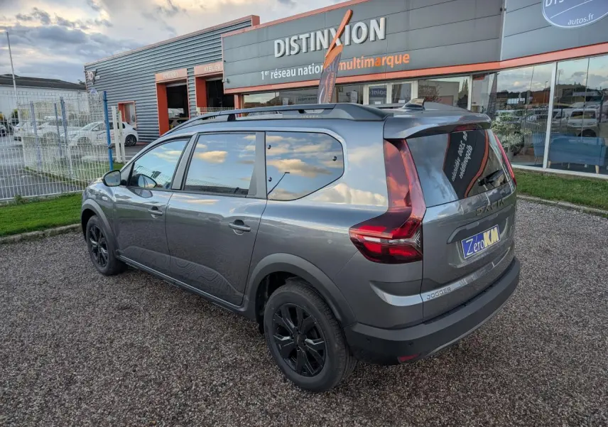Vue 3/4 arrière droite d'un Dacia Jogger gris métallisé avec jantes noires et barres de toit, stationné devant un garage.