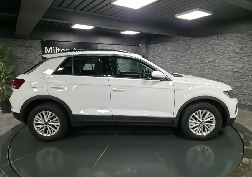 Volkswagen T-Roc blanc vu de profil côté gauche, avec jantes alliage et toit ouvrant en intérieur showroom.