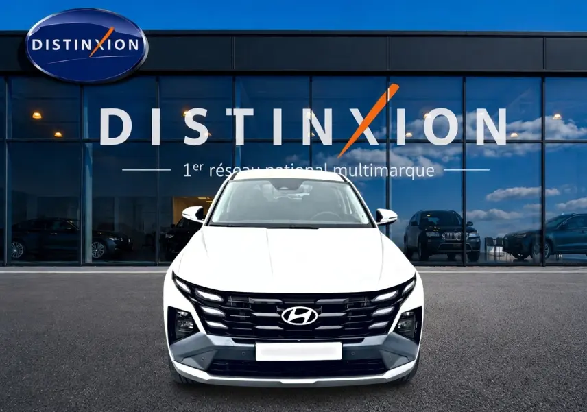 Vue avant d'un Hyundai Tucson blanc 2026 avec calandre noire distinctive, garé devant une vitrine de concession multimarque.