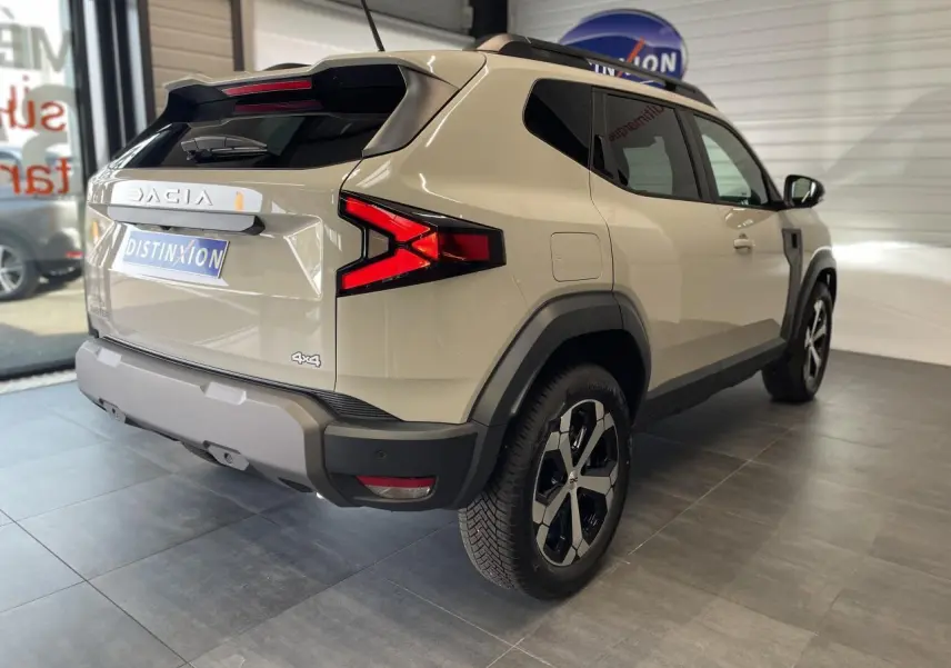 Vue 3/4 arrière droite du Dacia Duster beige 2025 avec jantes alliage noires et barres de toit noires.