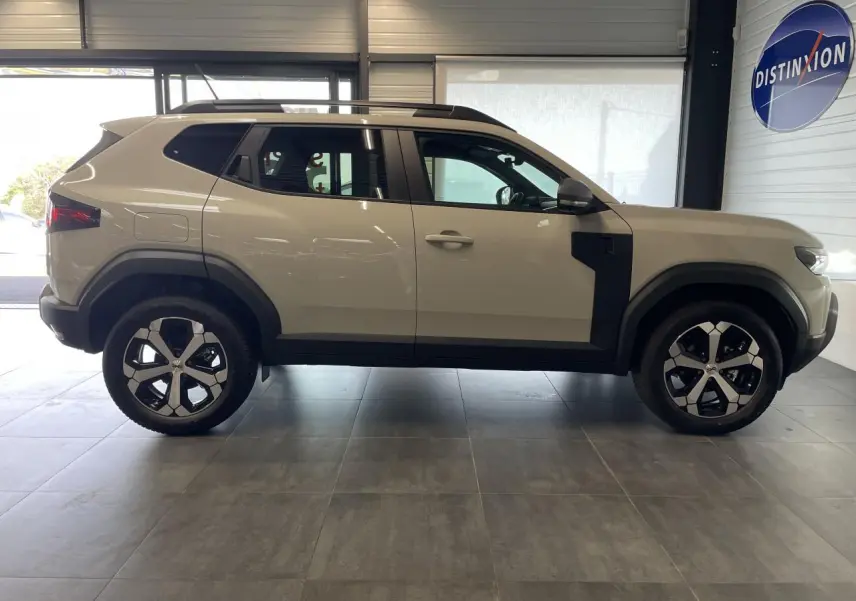 Vue latérale droite d'un Dacia Duster beige Sandstone avec jantes alliage 18 pouces et barres de toit noires.