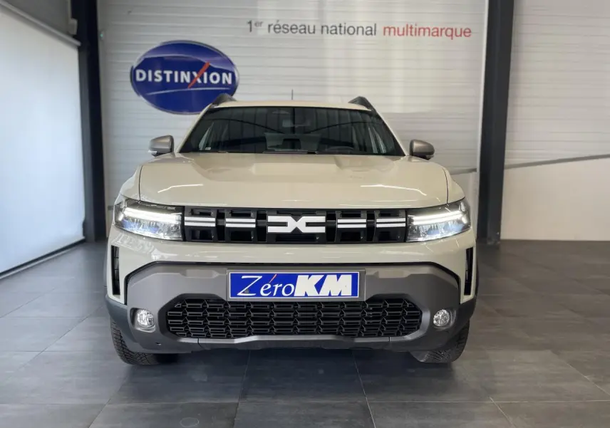 Vue avant d'un Dacia Duster beige 2025 avec calandre noire et feux LED allumés en intérieur showroom.