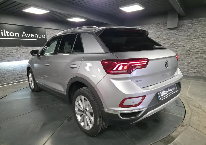 Vue 3/4 arrière droite d'un Volkswagen T-Roc gris métallisé avec feux arrière LED allumés en intérieur showroom.
