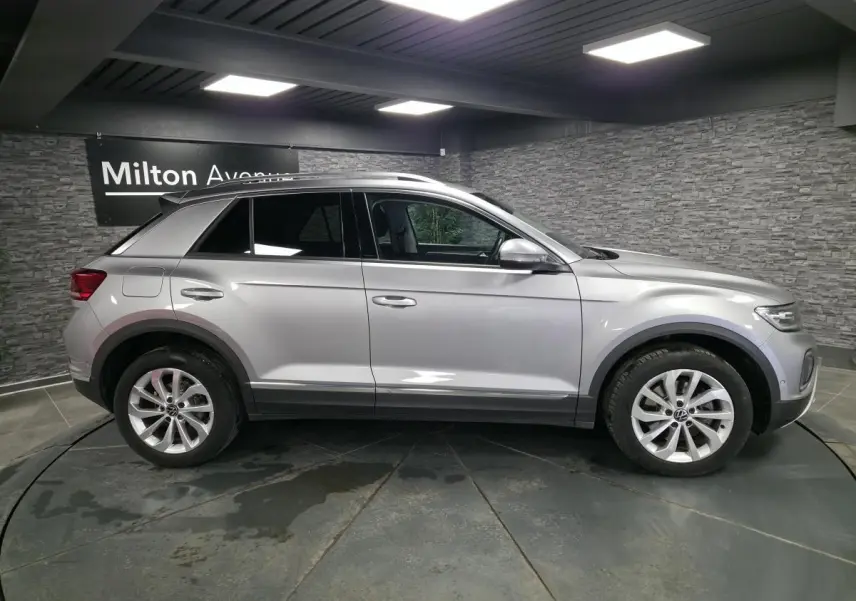 Vue latérale droite d'un Volkswagen T-Roc gris métallisé 2022, avec jantes alliage et barres de toit visibles.