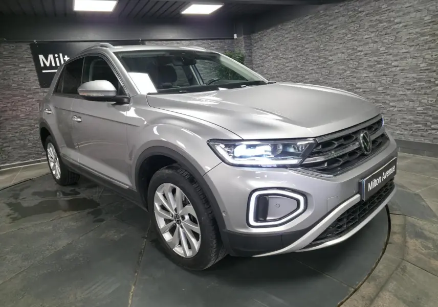 Volkswagen T-Roc gris métallisé vue 3/4 avant droit avec feux LED allumés en intérieur showroom.