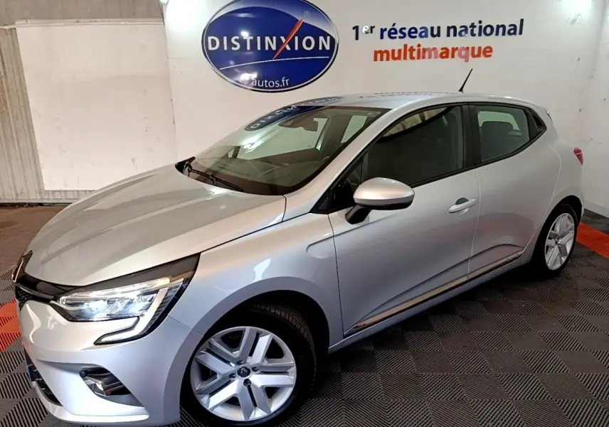 Vue 3/4 avant droite d'une Renault Clio gris clair avec feux LED allumés dans un showroom intérieur.