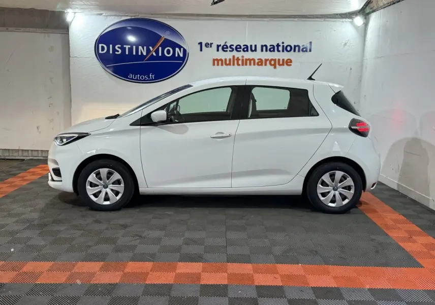 Profil droit d'une Renault ZOE blanche 2020 électrique, avec jantes simples et feux arrière allumés en intérieur showroom.