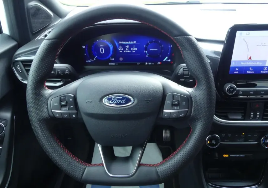 Vue intérieure frontale du volant noir perforé avec surpiqûres rouges du Ford Puma blanc, tableau de bord numérique et écran tactile central visibles.