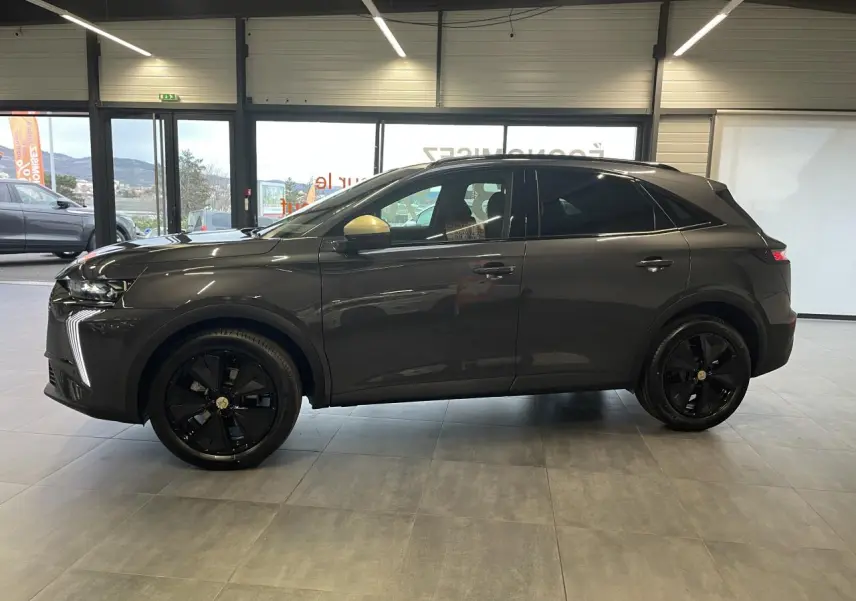 DS7 Crossback gris titanium en profil droit, jantes noires et rétroviseurs dorés satinés visibles en intérieur showroom.