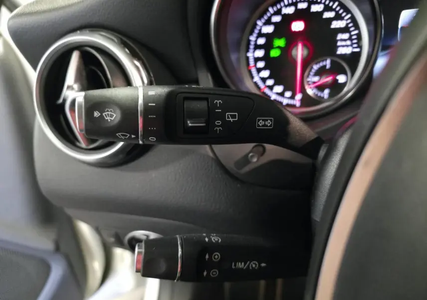 Détail du commodo gauche noir avec commandes d’essuie-glace et régulateur, tableau de bord partiellement visible d’une Mercedes GLA blanche.