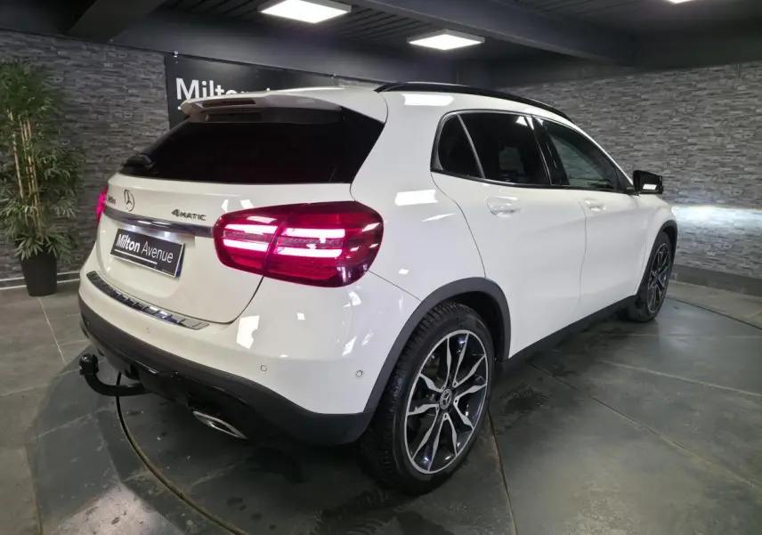 Vue 3/4 arrière droite d’un Mercedes GLA 220 d blanc avec jantes alliage 19’’ et attelage démontable en intérieur showroom.