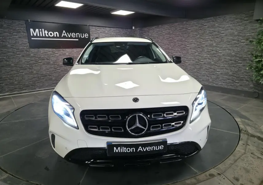 Vue avant d'un Mercedes GLA blanc avec calandre noire brillante et phares LED allumés dans un showroom.