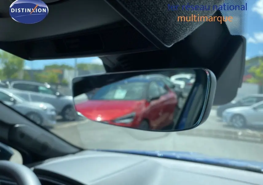 Rétroviseur intérieur montrant un reflet flou d’une voiture rouge, tableau de bord gris et ciel bleu visible.