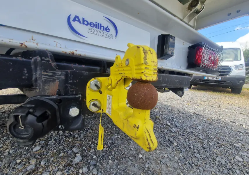 Gros plan sur l'attelage jaune et rouillé à l'arrière droit d'un Citroën Jumper blanc avec benne et ridelles grillagées.