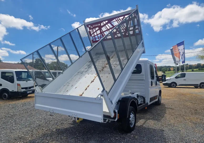 Vue 3/4 arrière droite du Citroën Jumper blanc avec benne relevée et ridelles grillagées sur fond de ciel bleu.