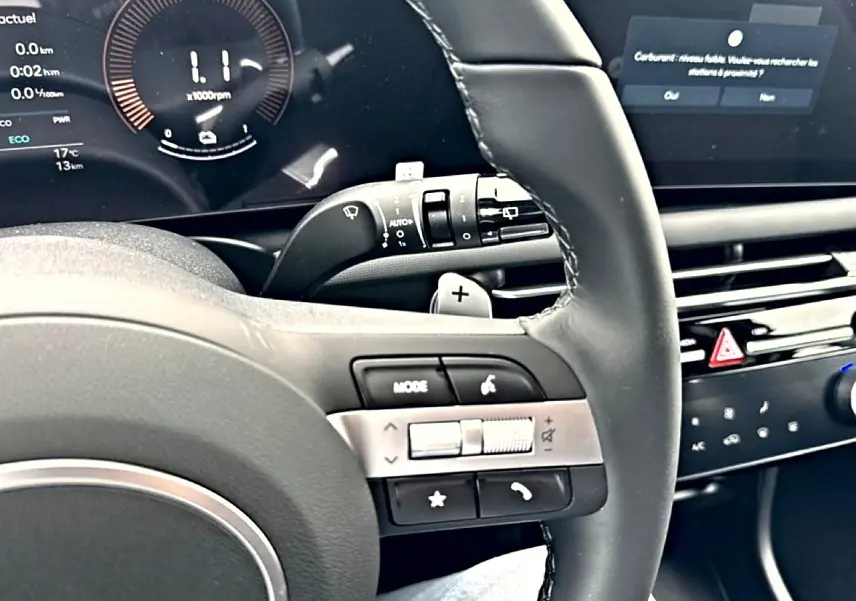 Vue rapprochée du volant cuir multifonction noir du Hyundai Tucson blanc, avec commandes audio et écran digital en arrière-plan.