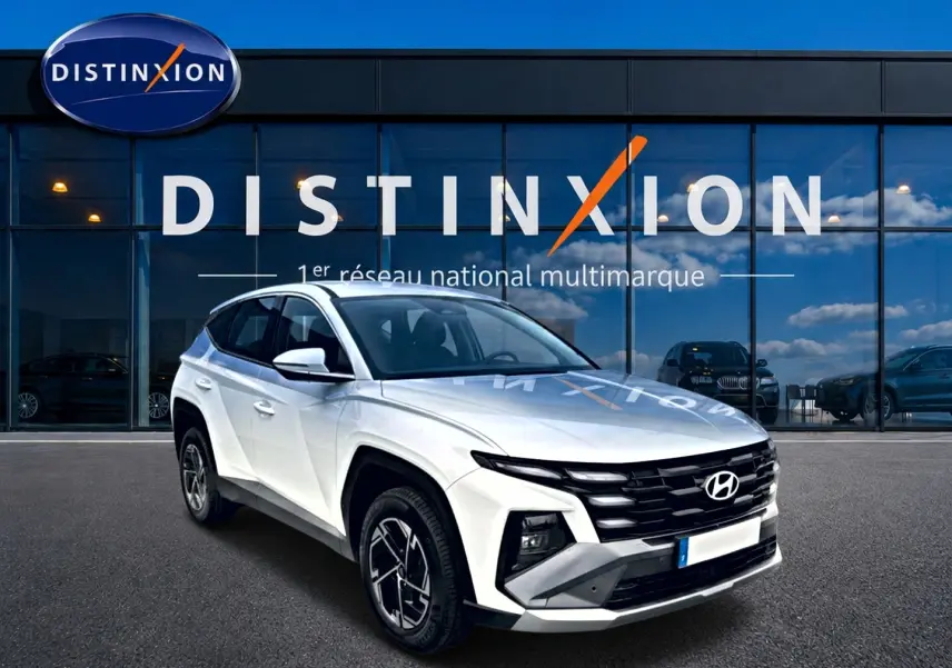 Vue 3/4 avant d'un Hyundai Tucson blanc 2026 avec calandre noire et jantes alliage 17 pouces devant un showroom Distinxion.