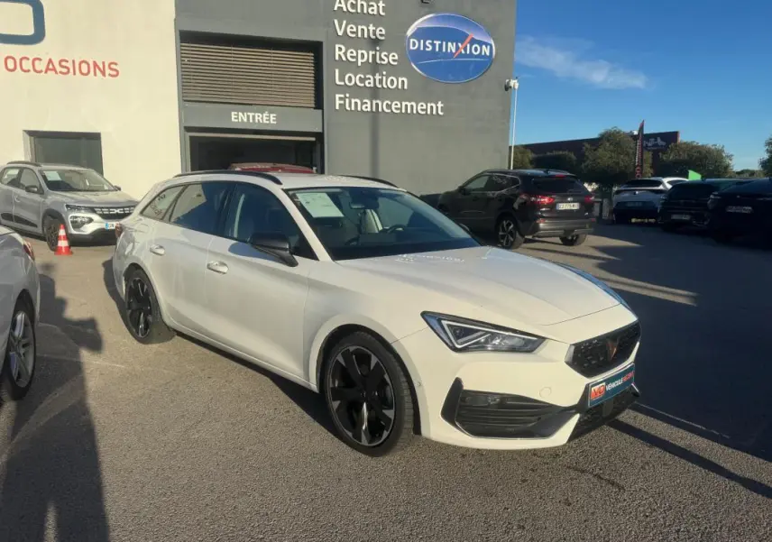 Vue 3/4 avant droite d'une CUPRA Leon Sportstourer blanche 2024 avec jantes noires, devant un concessionnaire.