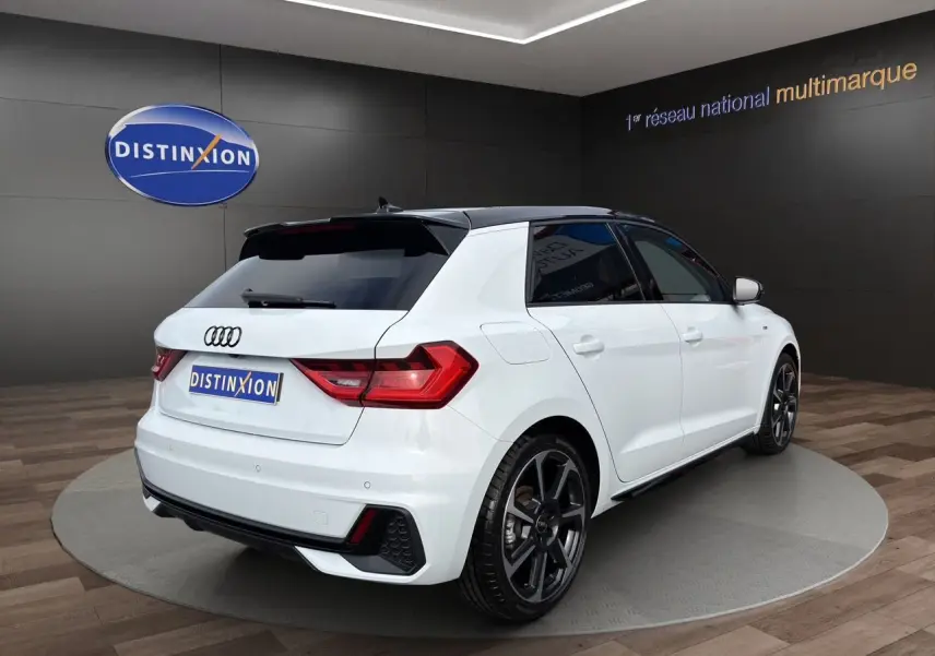Vue 3/4 arrière droite d'une Audi A1 Sportback blanche avec toit noir et jantes 18 pouces bicolores en hélice.