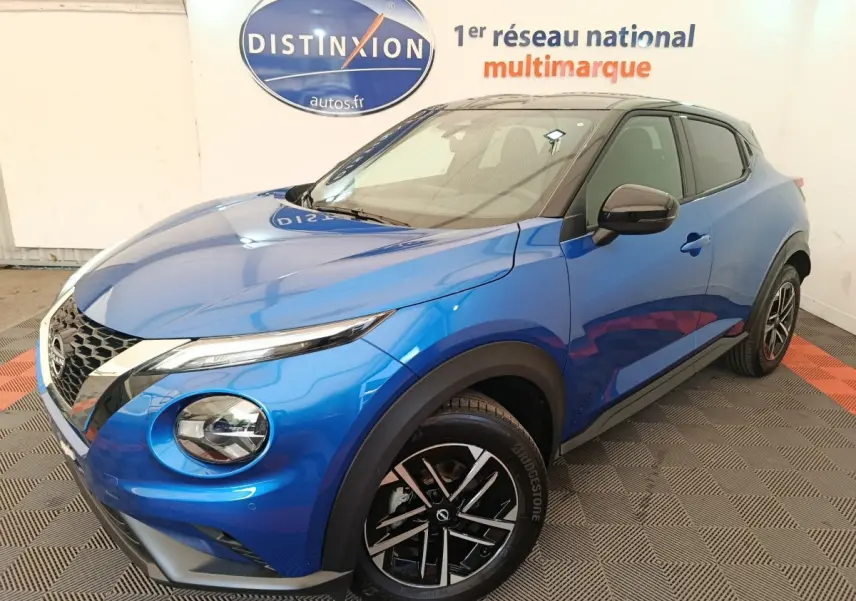 Nissan Juke bleu en 3/4 avant droit, avec toit noir et jantes alliage bicolores visibles en intérieur showroom.