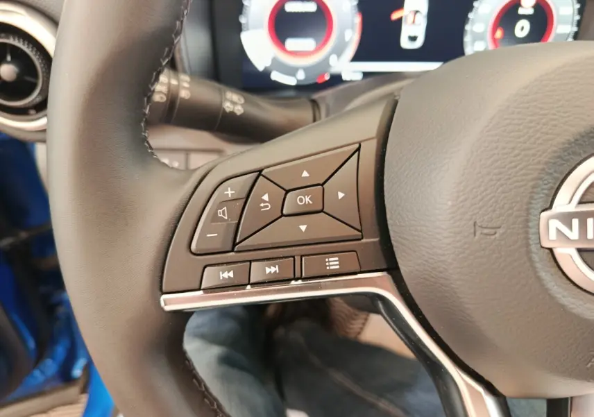 Gros plan sur les commandes gauche du volant cuir du Nissan Juke bleu 2025 avec tableau de bord numérique en arrière-plan.