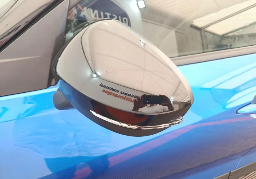 Rétroviseur chromé en gros plan côté gauche d’un Nissan Juke bleu 2025, reflet d’un intérieur d’atelier visible.