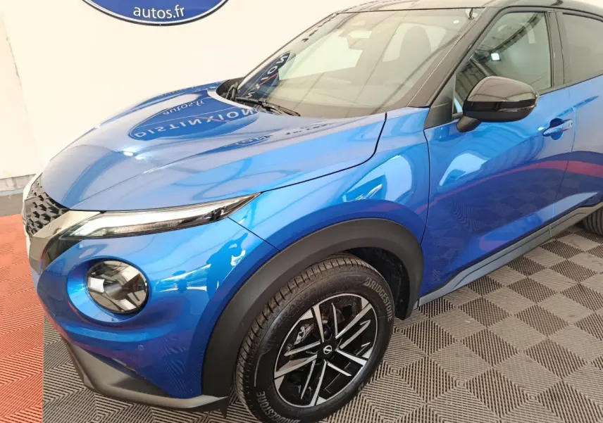 Nissan Juke bleu en 3/4 avant droit, avec phares ronds et jantes noires à motifs argentés.