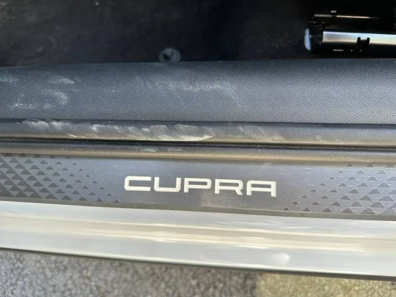 Gros plan sur la plaque de seuil de porte noire avec logo CUPRA blanc, sur un véhicule blanc Nevada métallisé.