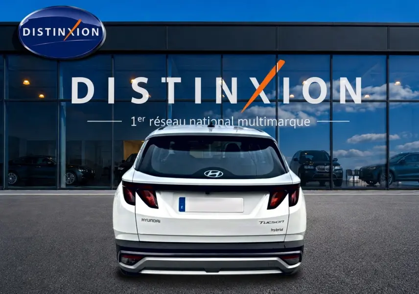 Vue arrière d'un Hyundai Tucson blanc hybride 2026, avec feux sombres et logo Hyundai visible sur un parking devant un bâtiment vitré.
