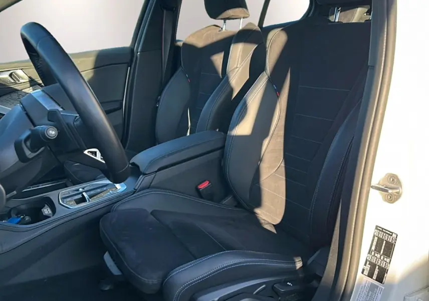 Vue intérieure côté conducteur d'une BMW Série 1 blanche, sièges sport en alcantara noir avec coutures bleues.