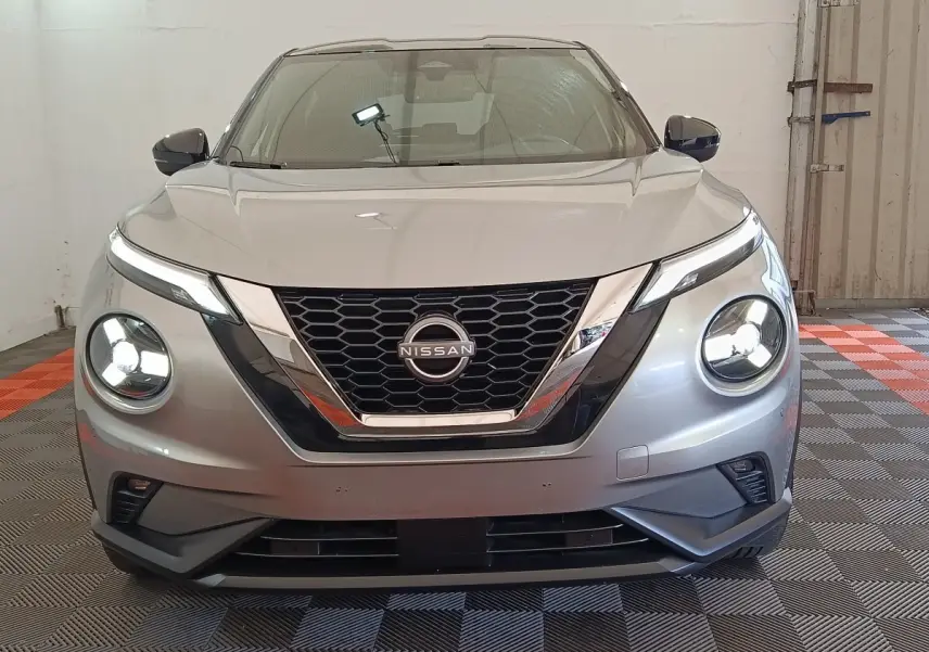 Vue frontale d'un Nissan Juke gris clair 2025 avec phares LED ronds allumés et calandre en nid d'abeille chromée.