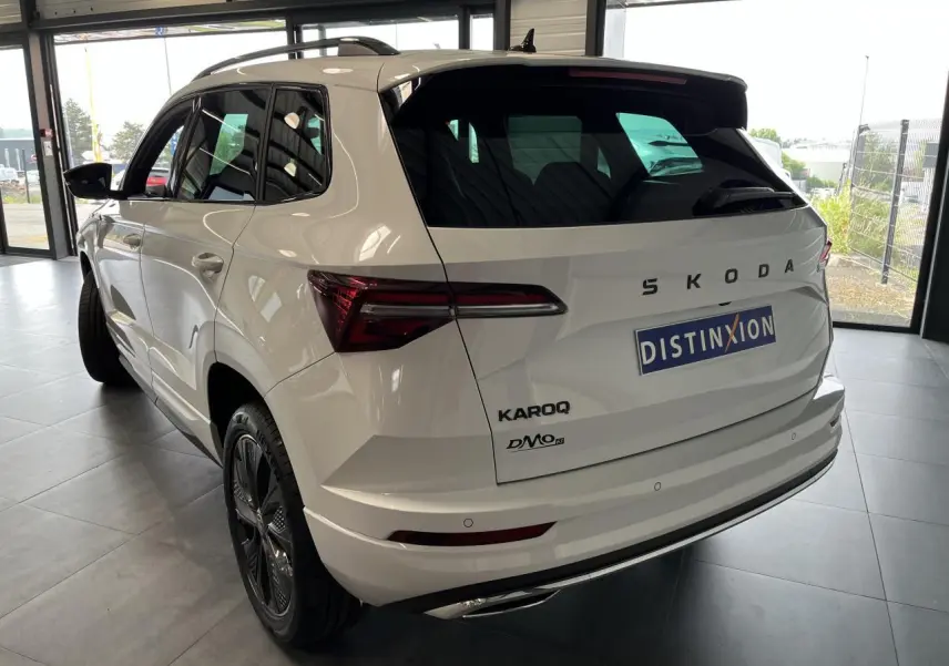 Vue 3/4 arrière droite du SUV blanc SKODA KAROQ 2025 avec jantes anthracite et logos noirs Sportline visibles.