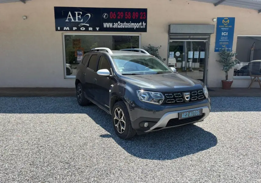 Dacia Duster gris en 3/4 avant droit, avec jantes alliage et barres de toit, stationné devant un garage AE2 Autos.