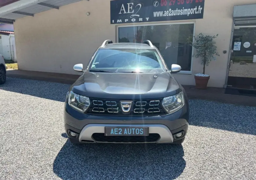 Vue avant d'un Dacia Duster gris 2021 Prestige avec calandre noire et barres de toit visibles, stationné devant un bâtiment.