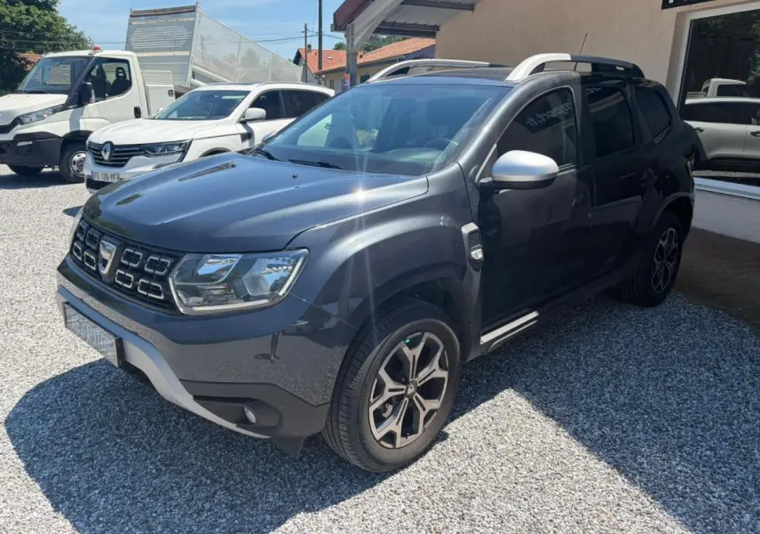 Dacia Duster gris vue 3/4 avant droit, avec jantes alliage et barres de toit visibles, stationné sur gravier.