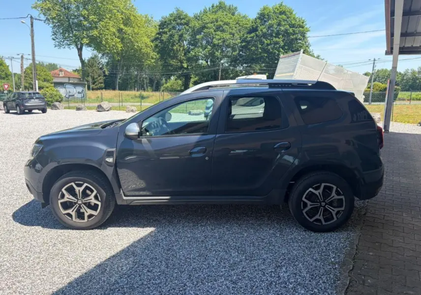 Vue de profil côté gauche d'un Dacia Duster gris 2021 avec jantes alliage et barres de toit noires.