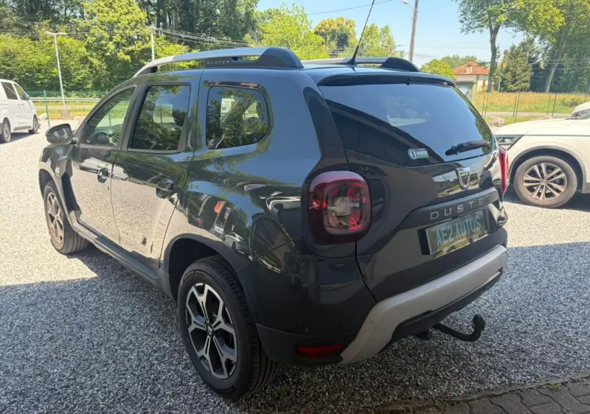 Vue 3/4 arrière droite d'un Dacia Duster gris 2021 avec barres de toit et attelage visible sur un sol gravillonné.