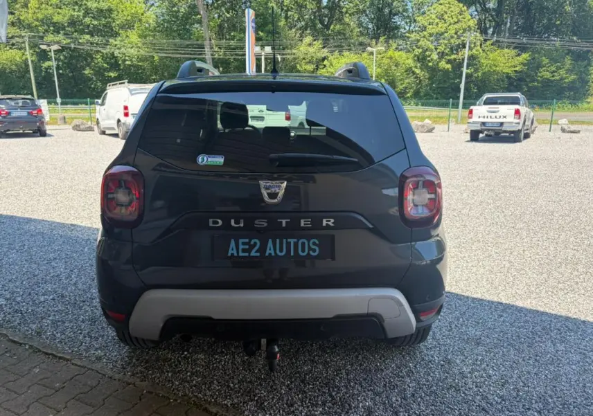 Vue arrière d'un Dacia Duster gris 2021 avec attelage, stationné sur un sol gravillonné en extérieur.