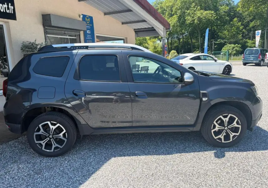 Vue de profil côté gauche d'un Dacia Duster gris 2021 avec jantes alliage et barres de toit argentées.