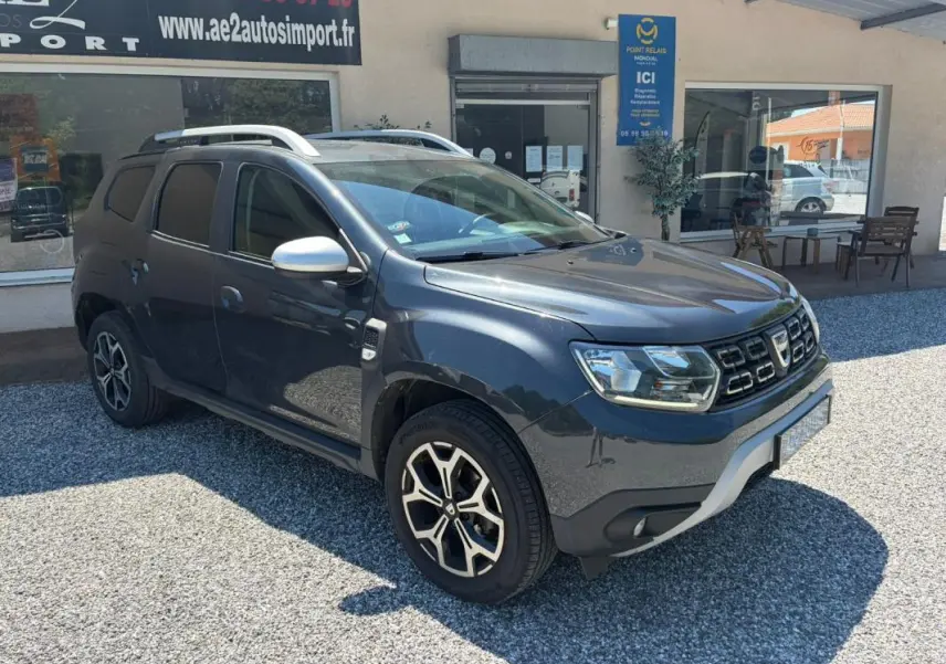 Dacia Duster gris en 3/4 avant droit, avec jantes alliage et barres de toit visibles, stationné devant un bâtiment.