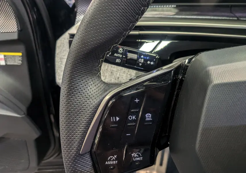 Gros plan sur le volant en cuir perforé noir du Peugeot 5008 Hybrid 145 GT, avec commandes multifonctions visibles.