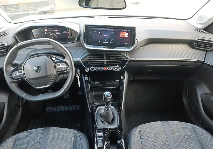 Intérieur avant de la Peugeot 208 1.2 100 Allure 2025, vue du conducteur sur tableau de bord et levier de vitesses manuel.