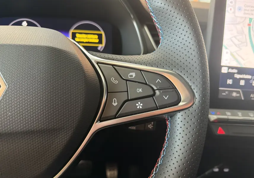 Gros plan sur la partie droite du volant perforé avec surpiqûres bleues et rouges du Renault Captur E-Tech 160 Esprit Alpine, tableau de bord numérique visible.