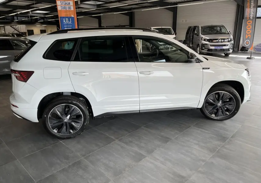 Vue latérale droite d'un SUV Skoda Karoq blanc 2025 avec jantes alliage noires et badges Sportline visibles.
